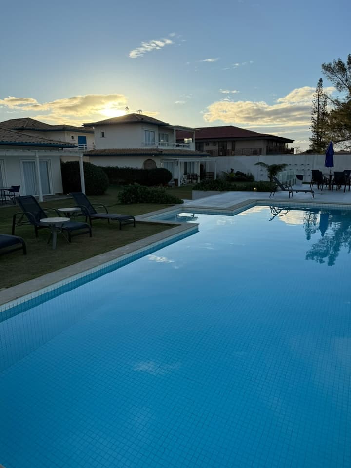 Piscina ampla de água azul e calma ao entardecer, com espreguiçadeiras no gramado e casas brancas de telhado colonial ao fundo.