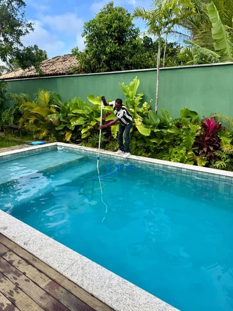 Profissional em pé na borda aspira o fundo de uma piscina de água azul, diante de um jardim tropical.